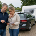 Echtpaar staat naast een auto met caravan en bekijkt de kosten in een rustige Nederlandse kampeeromgeving.