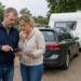 Echtpaar staat naast een auto met caravan en bekijkt de kosten in een rustige Nederlandse kampeeromgeving.